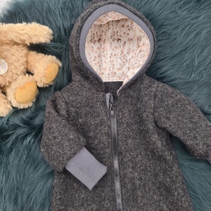 Könnte beinhalten: Eine dunkelgraue Kapuzenjacke mit durchgehendem Reißverschluss, grauen Bündchen und einem Innenfutter mit floralem Muster. Die Jacke wird auf einer teal-farbenen, pelzigen Oberfläche mit einem Teddybären im Hintergrund gezeigt.