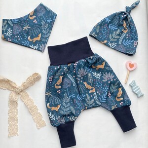 Puede incluir: Un conjunto de ropa para bebé con un babero, pantalones y gorro con estampado azul y blanco. El estampado presenta zorros, hojas y flores.