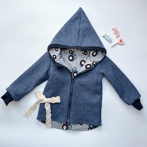 Könnte beinhalten: Eine blaue Kapuzenjacke mit Reißverschluss und Traktor-Muster-Futter. Die Jacke hat eine weiße Spitzen-Schleife und schwarze Bündchen.