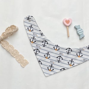 Op de afbeelding: Een baby slabbetje met een nautisch thema, met grijze en witte strepen en zwarte ankerontwerpen met oranje accenten. Op het slabbetje staan de woorden "MOIN" en "AHOI". Een crème-kleurige kanten strik en houten speelgoed zijn ook in de afbeelding.