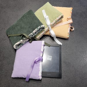 Peut inclure: Quatre étuis pour liseuse en tissu côtelé vert, jaune et lavande, avec bordures à motifs floraux et fermetures à ruban. Une liseuse noire avec le texte "fnac" et "kobo" est partiellement visible.