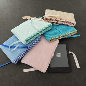 Peut inclure: Une collection de cinq housses rectangulaires en tissu pour liseuse électronique dans des tons pastel incluant vert menthe, bleu clair, rose, beige et bleu sarcelle. Chaque housse a un ruban de fermeture et une doublure à motifs. Une liseuse noire.