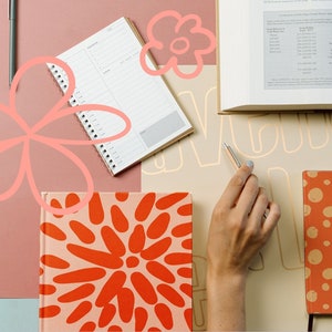 May include: A flat lay of a desk with various notebooks, pens, and a book. The notebooks have different patterns, including orange and red polka dots, and a floral design. The book is open to a page with a table of data. The desk is covered in a light brown surface.