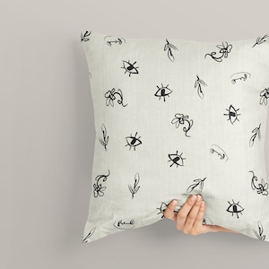 May include: A white pillow with a black and white pattern of eyes, leaves, and flowers. The pillow is being held by a hand.