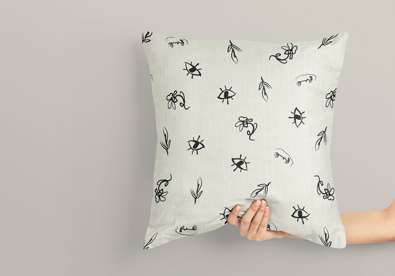 May include: A white pillow with a black and white pattern of eyes, leaves, and flowers. The pillow is being held by a hand.
