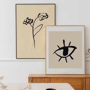 May include: Two framed abstract art prints on a wooden shelf. The print on the left features a black line drawing of a flower on a beige background. The print on the right features a black line drawing of an eye on a beige background.