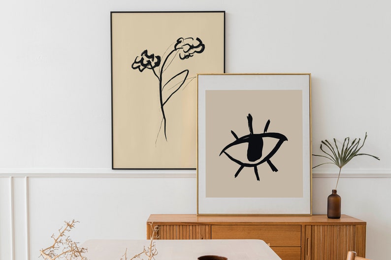 May include: Two framed abstract art prints on a wooden shelf. The print on the left features a black line drawing of a flower on a beige background. The print on the right features a black line drawing of an eye on a beige background.