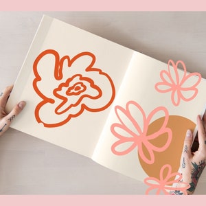 May include: An open sketchbook with hand-drawn illustrations of flowers in orange and pink. The sketchbook is being held by a person with tattoos on their arms.