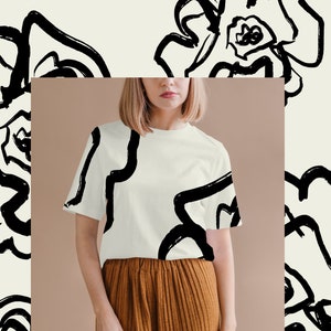 May include: A white t-shirt with a black abstract floral print. The shirt is being modeled by a person with short blonde hair. The background is a light brown color.
