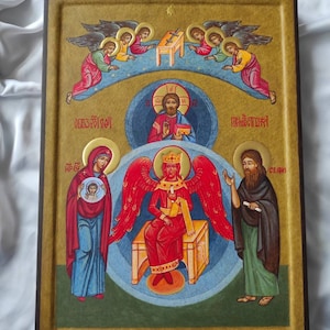 Op de afbeelding: Een religieus icoon dat de Annunciatie voorstelt. De centrale figuur is de aartsengel Gabriël, met vleugels, een kroon en een gouden staf. Hij zit op een troon en is omringd door een blauwe halo. De Maagd Maria staat links, ze houdt een klein icoon van de baby Jezus vast. De Heilige Geest wordt voorgesteld door een duif boven de aartsengel Gabriël. Het icoon is geschilderd op een houten paneel en ingelijst in een donkerhouten lijst.