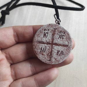 May include: A white pendant with a brown cross and the letters "XC", "IC", "KA", and "NI" inscribed on it. The pendant is on a black leather cord.