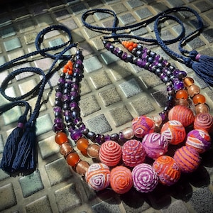 Puede incluir: Un collar artesanal con una combinación de cuentas moradas, naranjas y transparentes, junto con cuentas texturizadas, estampadas y envueltas en hilo de colores. El collar tiene un cordón trenzado azul marino con borlas.