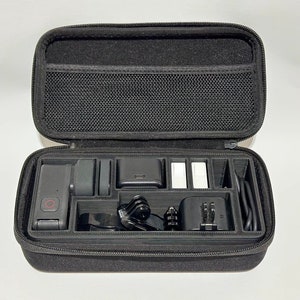 May include: Black zippered carrying case, open to reveal a camera and accessories. The case has a mesh pocket in the lid and compartments for various items, including a camera, batteries, and charging cables. The case is designed to protect and organize camera equipment.