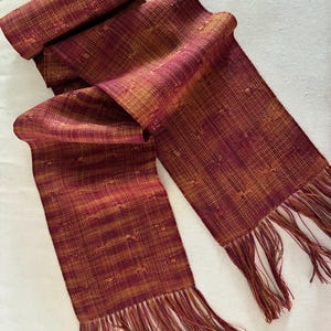 Pu&ograve; includere: Una sciarpa tessuta bordeaux e oro, con un motivo strutturato e bordi sfrangiati. La sciarpa &egrave; drappeggiata, mostrando la sua lunghezza e l'interazione dei colori. Il tessuto appare morbido e probabilmente &egrave; fatto di fibre naturali.
