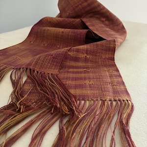 Pu&ograve; includere: Sciarpa tessuta bordeaux e oro con bordo a frange. La sciarpa &egrave; drappeggiata, mostrando la sua consistenza e le variazioni di colore. La frangia &egrave; un mix di fili bordeaux e oro.