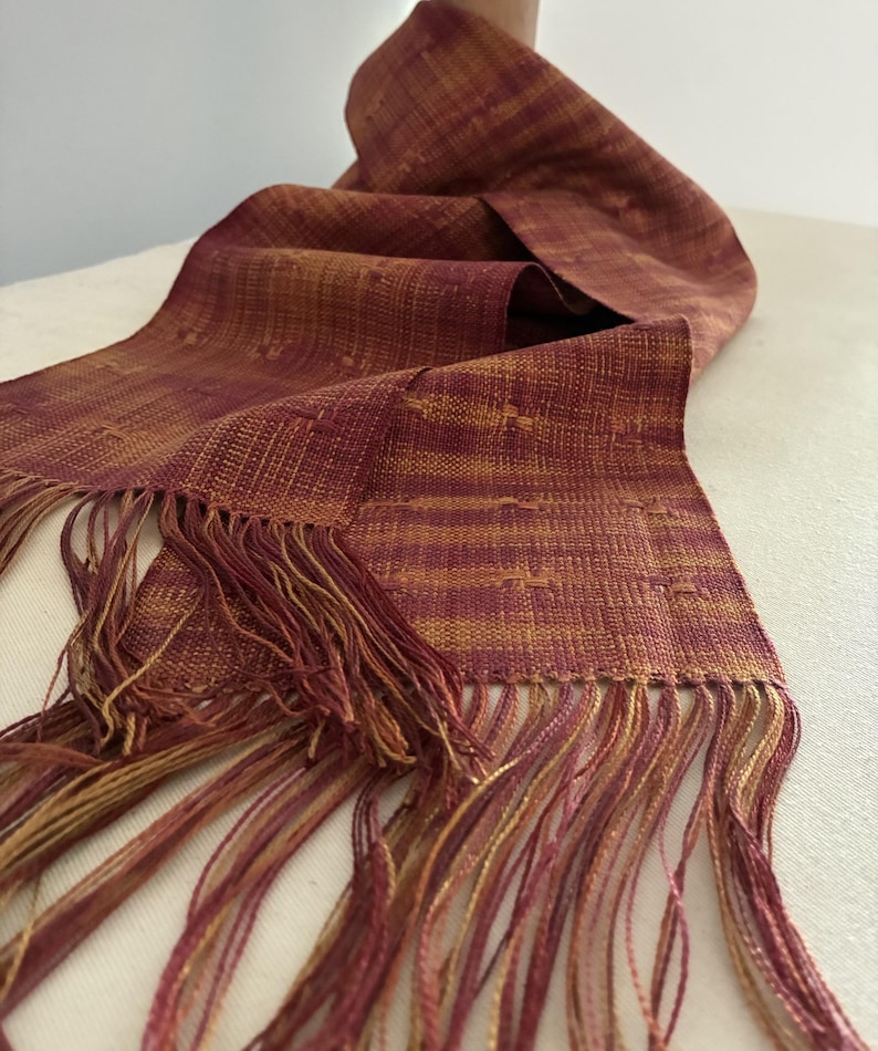 Pu&ograve; includere: Sciarpa tessuta bordeaux e oro con bordo a frange. La sciarpa &egrave; drappeggiata, mostrando la sua consistenza e le variazioni di colore. La frangia &egrave; un mix di fili bordeaux e oro.