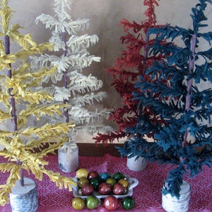 May include: Four felt Christmas trees in different colors: yellow, white, red, and blue. The trees are standing on a table with a red and white patterned tablecloth. A bowl of colorful ornaments is in front of the trees.