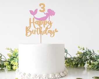 May include: Gold glitter cake topper with the words "Happy Birthday" and a pink glitter mermaid with a tail and the number "3".
