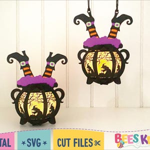 May include: Two black witch legs dangle from a hanging cauldron with a purple rim. The cauldron has a yellow glow inside and a silhouette of a witch stirring a pot. The background is a white wood panel.