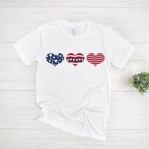 May include: White t-shirt with three red, white, and blue hearts in a row. The hearts are patterned with stars and stripes.