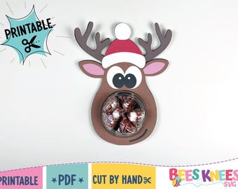 Peut inclure: Un porte-bonbons en forme de renne marron avec un bonnet de Père Noël rouge et blanc. Le support contient un bol transparent rempli de bonbons emballés. L'image comprend le texte "PRINTABLE" et "CUT BY HAND".