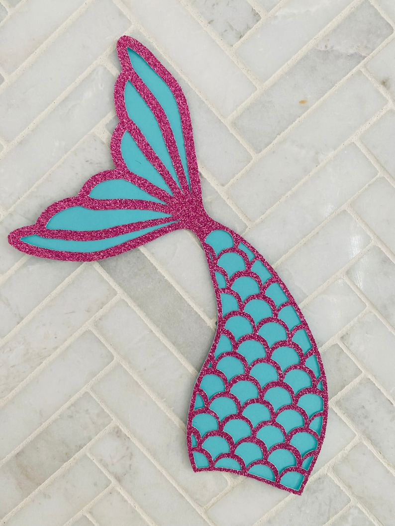 May include: A turquoise mermaid tail cutout with pink glitter. The tail has a scale pattern and a fin.