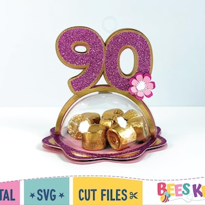 May include: A decorative 90th birthday-themed candy holder. The number 90 is in glittery purple with a gold outline, and a pink flower is attached. The clear dome contains gold-wrapped candies, all set on a scalloped base.