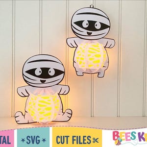 May include: Two white paper mummy cutouts with yellow and white patterned paper lanterns inside. The mummies have black eyes and mouths. The cutouts are hanging on a white wood panel background.