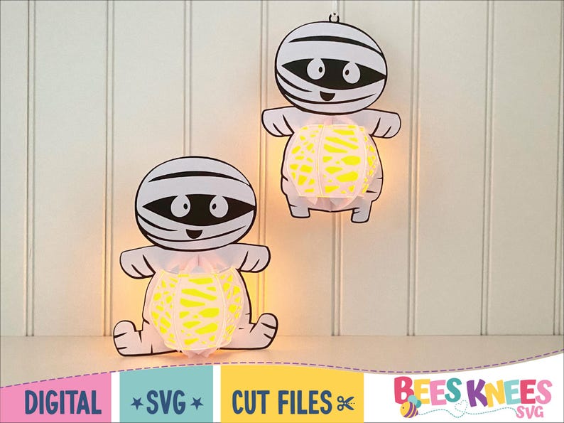 May include: Two white paper mummy cutouts with yellow and white patterned paper lanterns inside. The mummies have black eyes and mouths. The cutouts are hanging on a white wood panel background.