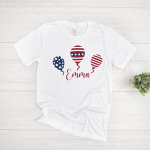 May include: White t-shirt with a red, white, and blue balloon graphic and the name "Emma" printed on the front.