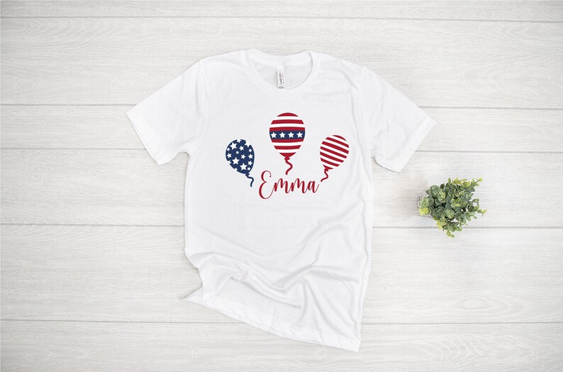 May include: White t-shirt with a red, white, and blue balloon graphic and the name "Emma" printed on the front.
