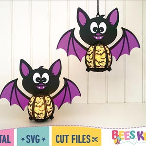 May include: Two purple and black bat shaped Halloween decorations with a yellow and black patterned lantern inside. The bats are hanging from a white background.
