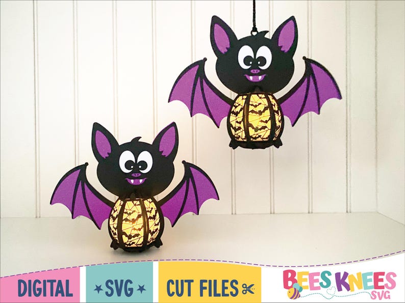 May include: Two purple and black bat shaped Halloween decorations with a yellow and black patterned lantern inside. The bats are hanging from a white background.