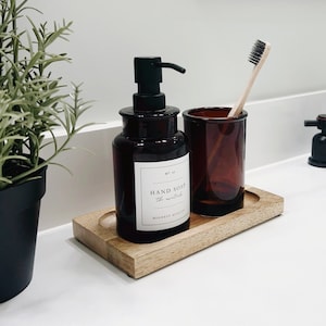 以下が含まれることがあります： 「HAND SOAP」と書かれた茶色の石鹸ディスペンサー、茶色のガラスのコップ、歯ブラシ、黒い蛇口がついたバスルームの洗面台です。