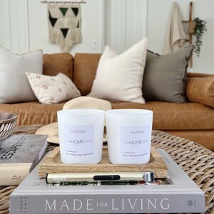 以下が含まれることがあります： ラベルに「Nostalgia theHOMEmind」と「sandalwood 8.5 oz / 100% Soy Wax / Hand Poured」と書かれた白いキャンドルが2本。キャンドルは木製トレイの上に置かれ、トレイの上には「MADE FOR LIVING」と書かれた本の横にライターが置かれている。