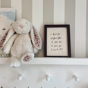 May include: A cream-coloured plush rabbit with floral-patterned inner ears and foot pads, positioned next to a dark brown wooden frame containing a white fabric insert embroidered with the repeated black text "I love you" five times.