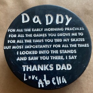 May include: Black hockey puck with white text that reads "DADDY" and a message of thanks to a dad from a child. The message reads "FOR ALL THE EARLY MORNING PRACTICES FOR ALL THE GAMES YOU DROVE ME TO FOR ALL THE TIMES YOU TIED MY SKATES BUT MOST IMPORTANTLY FOR ALL THE TIMES I LOOKED INTO THE STANDS AND SAW YOU THERE, I SAY THANKS DAD Love Abella".