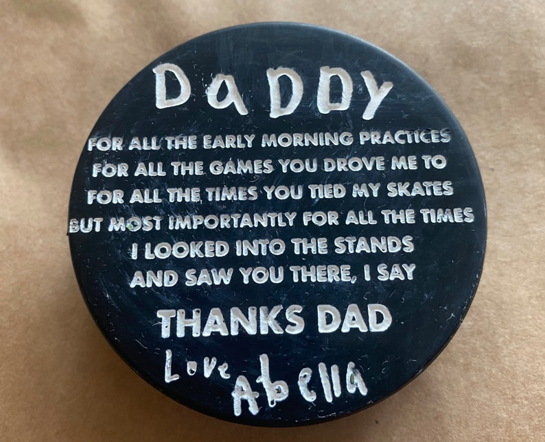 May include: Black hockey puck with white text that reads "DADDY" and a message of thanks to a dad from a child. The message reads "FOR ALL THE EARLY MORNING PRACTICES FOR ALL THE GAMES YOU DROVE ME TO FOR ALL THE TIMES YOU TIED MY SKATES BUT MOST IMPORTANTLY FOR ALL THE TIMES I LOOKED INTO THE STANDS AND SAW YOU THERE, I SAY THANKS DAD Love Abella".