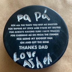 May include: Black hockey puck with white text that reads "pa P&aacute; FOR ALL THE TIMES YOU GOT ME DRESSED FOR TAPING MY STICK AND TYING MY SKATES FOR ALWAYS MAKING SURE I HAVE SNACKS FOR CHEERING ME ON FROM THE STANDS FOR BEING MY BIGGEST FAN ON AND OFF THE RINK THANKS DAD LOVE Ash"
