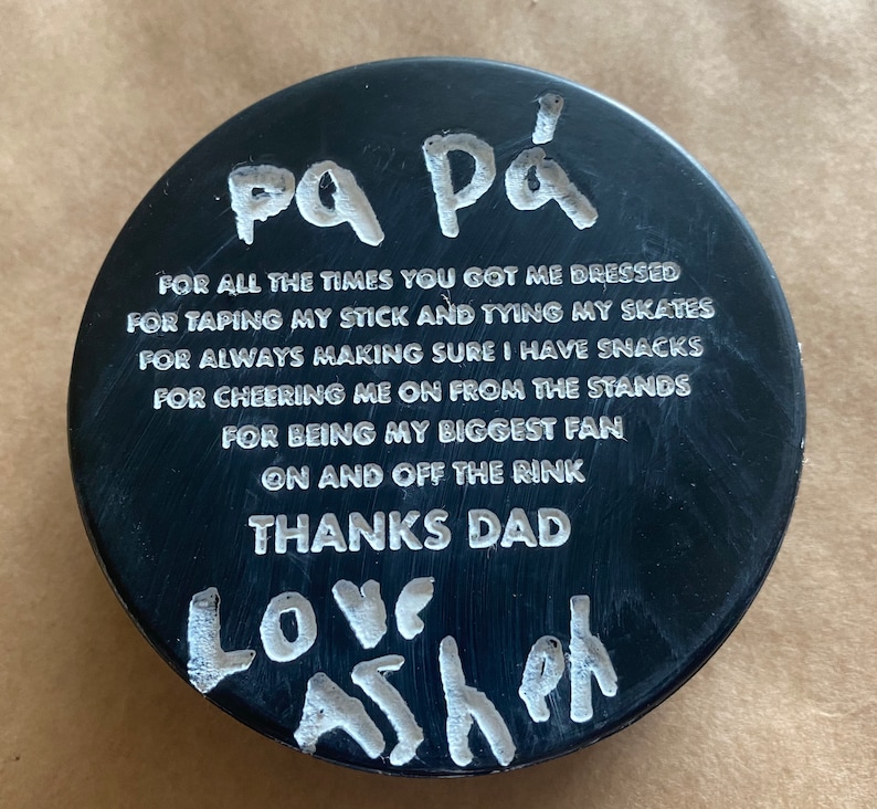 May include: Black hockey puck with white text that reads "pa P&aacute; FOR ALL THE TIMES YOU GOT ME DRESSED FOR TAPING MY STICK AND TYING MY SKATES FOR ALWAYS MAKING SURE I HAVE SNACKS FOR CHEERING ME ON FROM THE STANDS FOR BEING MY BIGGEST FAN ON AND OFF THE RINK THANKS DAD LOVE Ash"
