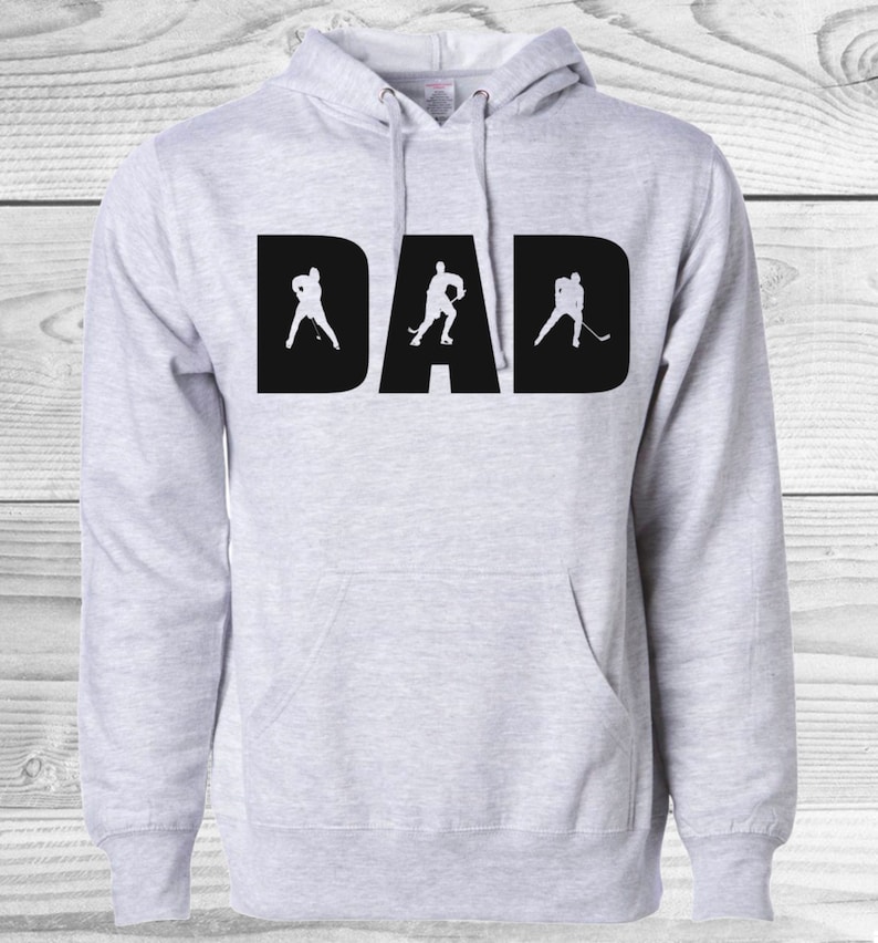 May include: Light gray hoodie with the word "DAD" in large black letters. The letter shapes incorporate hockey player silhouettes. The hoodie has a front pocket and a drawstring hood.