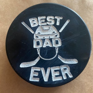May include: Black hockey puck with a white design that says "Best Dad Ever" with a hockey helmet and two hockey sticks.