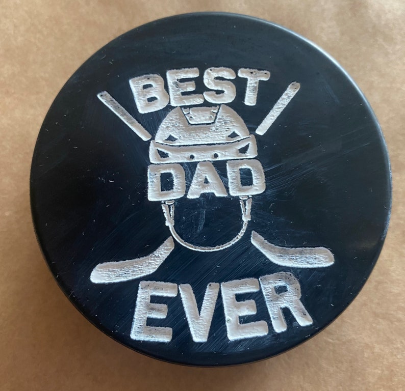 May include: Black hockey puck with a white design that says "Best Dad Ever" with a hockey helmet and two hockey sticks.
