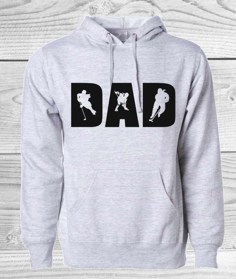May include: Light gray hoodie with the word "DAD" in large black letters. The letters are filled with white silhouettes of hockey players in action. Features a hood and a front pocket.