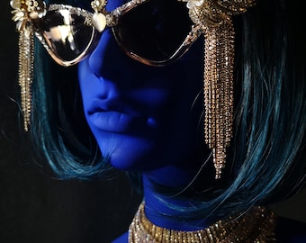 Puede incluir: Un maniquí azul que lleva un collar de cuentas doradas y unas gafas de sol doradas con adornos de cristal y flecos colgantes.