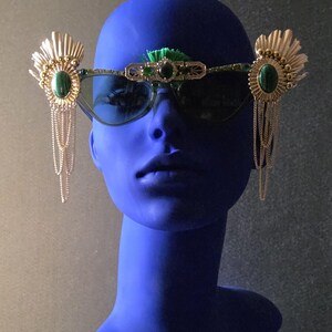 May include: Green-tinted sunglasses with gold-coloured decorative elements and green gemstones. The design features dangling chains and a decorative brow bar, creating a unique and artistic accessory.
