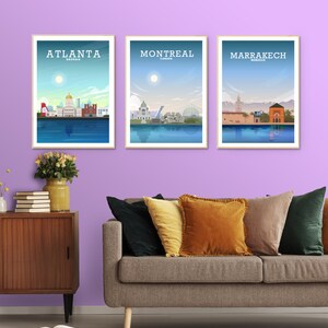 May include: Three framed city skyline posters with a light blue background and white text. The posters are titled "Atlanta, Georgia", "Montreal, Canada", and "Marrakech, Morocco". The posters are hanging on a light purple wall above a gray couch with pillows.