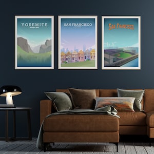 May include: Three framed posters on a dark blue wall above a brown leather couch. The posters are of Yosemite National Park, San Francisco, California, and San Francisco, California. The poster of Yosemite shows a mountain range with a valley in the foreground. The poster of San Francisco shows a row of colorful Victorian houses. The poster of San Francisco shows a baseball stadium.