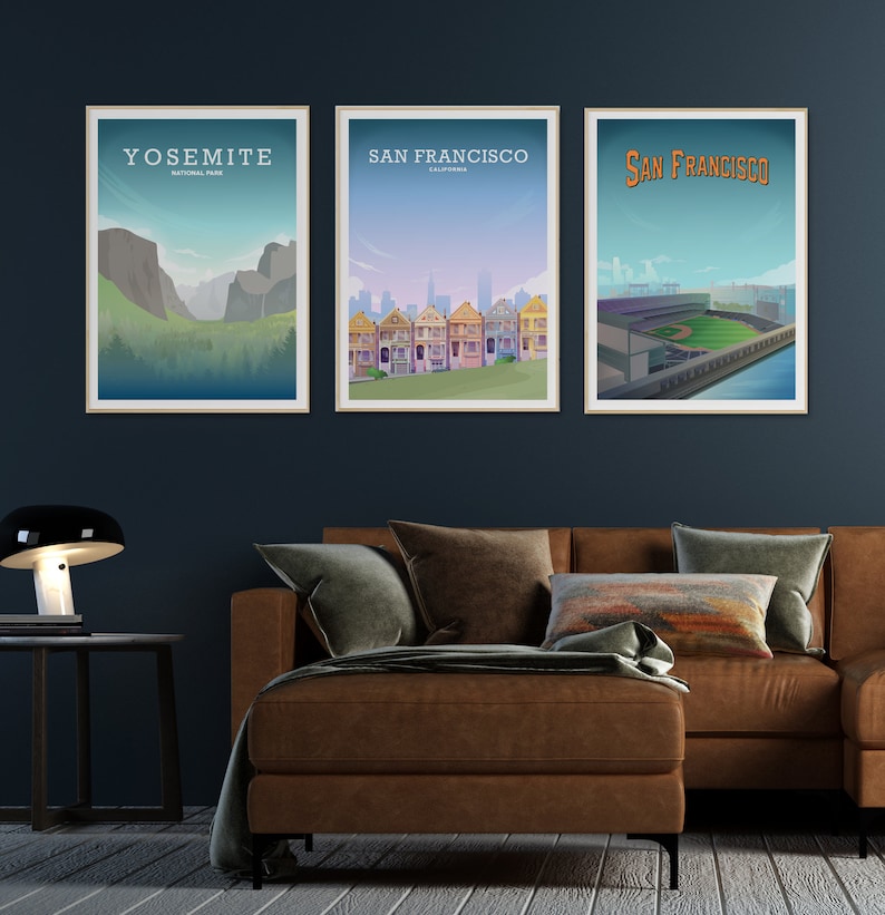 May include: Three framed posters on a dark blue wall above a brown leather couch. The posters are of Yosemite National Park, San Francisco, California, and San Francisco, California. The poster of Yosemite shows a mountain range with a valley in the foreground. The poster of San Francisco shows a row of colorful Victorian houses. The poster of San Francisco shows a baseball stadium.