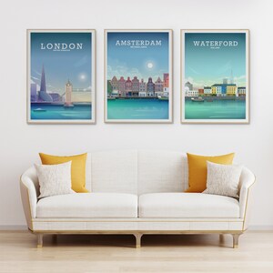 May include: Three framed travel art prints featuring cityscapes of London, Amsterdam, and Waterford. Each print has a gold frame and depicts iconic landmarks and buildings in a stylized, colorful illustration. The prints are displayed above a white sofa with yellow and white pillows.